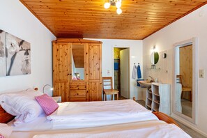 A bedroom with a wooden ceiling, a bed with white bedding, a wooden wardrobe, a bathroom with a sink and mirror, and a door leading to another room.
