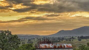A rustic, single-story building with a corrugated metal roof, surrounded by a fence and greenery, set against a backdrop of a cloudy sky and distant hills.