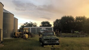 Paisaje rural con una camioneta verde estacionada en un campo cubierto de hierba, un silo y un edificio al fondo.