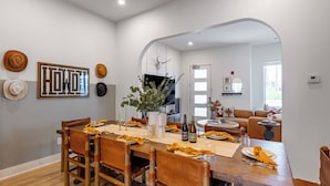 Un comedor con una mesa de madera dispuesta para cuatro personas, rodeada de sillas. Hay sombreros en la pared, un gran adorno de palabras enmarcado y una planta sobre la mesa.