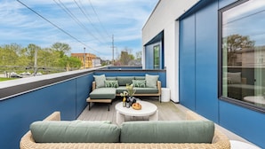 Un área de estar exterior moderna con un sofá y sillones, una mesa redonda y vista a la calle.