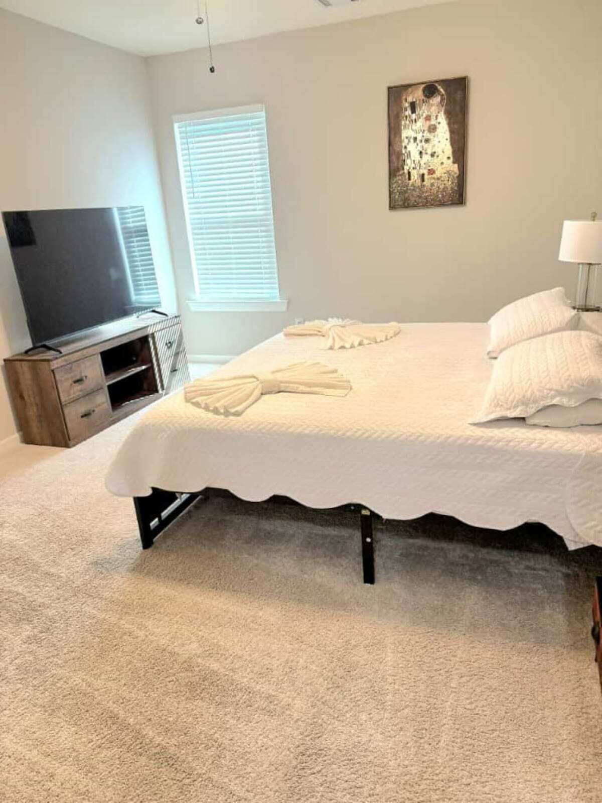 Un dormitorio con cama, televisión, un cuadro y ventana con persianas.