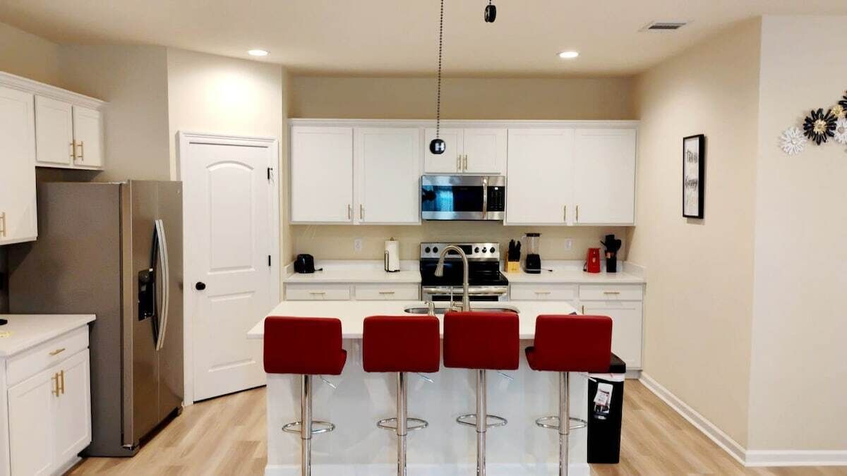 Una cocina moderna con armarios blancos, un refrigerador de acero inoxidable, un microondas y una estufa a gas. Hay cuatro taburetes rojos y un bote de basura negro.