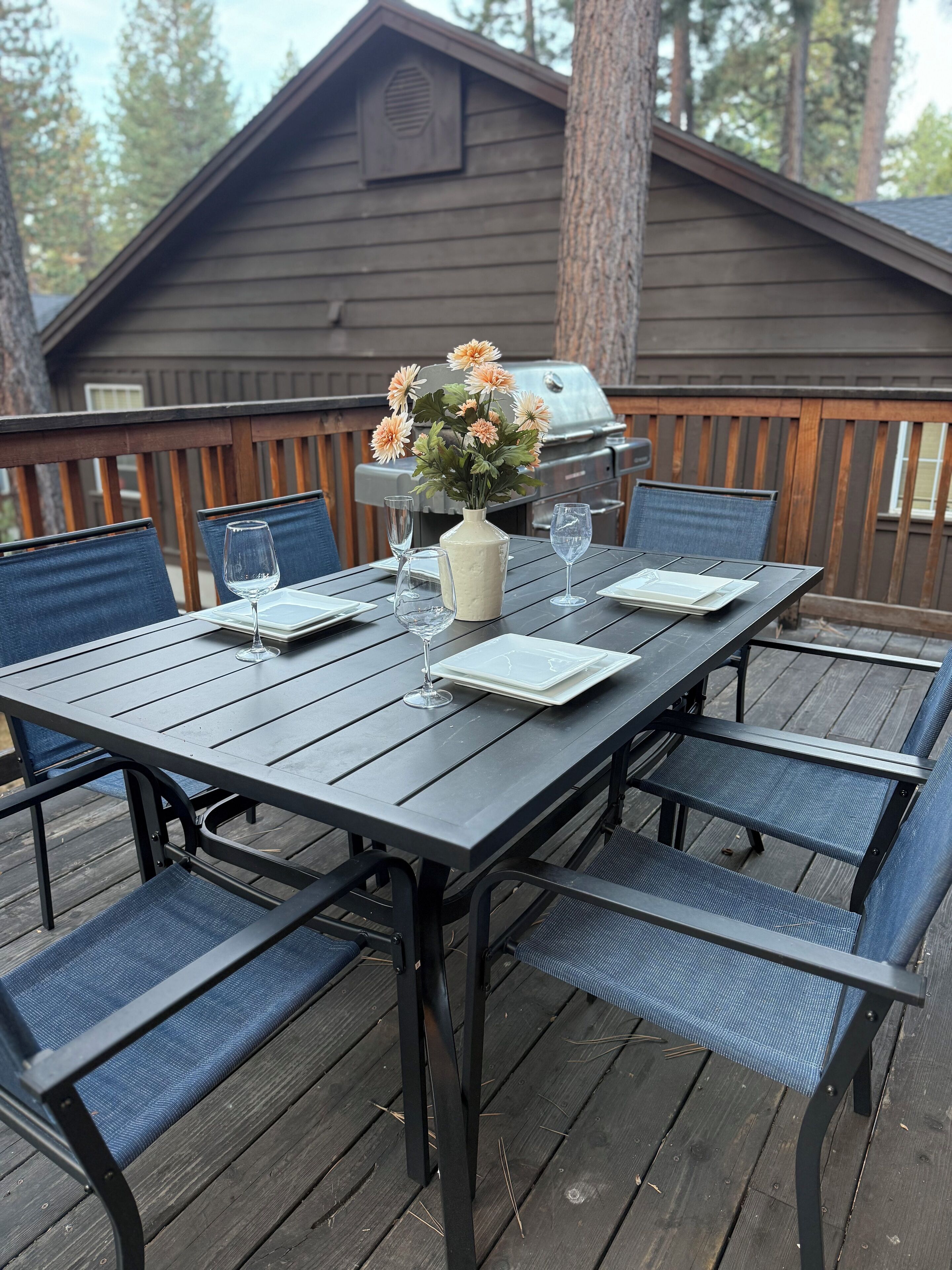 A deck with a table set for dining, chairs, a grill, and a wooden cabin in the background.