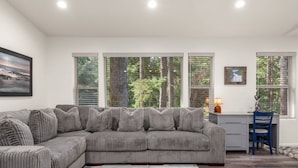A modern living room with a grey sectional sofa, a desk with a chair, and large windows with blinds.