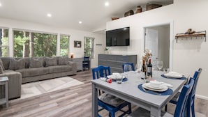 A modern living room with a grey sofa, a dining table set for four, and a TV mounted on the wall.
