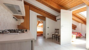 Een keuken met een gasfornuis, een spoelbak en een raam boven het fornuis. Er staat een stoel in de ruimte en een deur naar een andere kamer.
