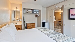 Een hotelkamer met een bed, een houten dressoir, een spiegel, een plafondventilator, een kleine koelkast en een deur naar een eethoek.