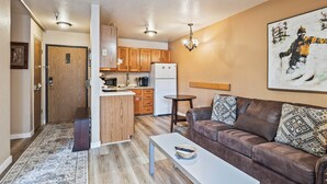 Een gezellig, compacte ruimte met een kitchenette, een bruine leren bank, een witte salontafel en een schilderij aan de muur.