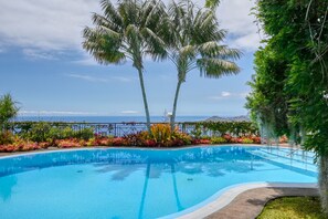 Une piscine bordée de palmiers et agrémentée de plantes aux couleurs vives, entourée d’une clôture et offrant une vue sur l’océan.