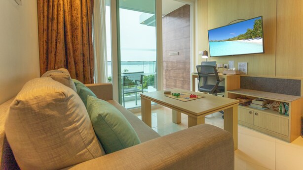 A modern living room with a sofa, a coffee table, a flat-screen TV, and a view of the outdoors through a glass door. - Sea View Beach Bar Beach Front c114 (Rawai)