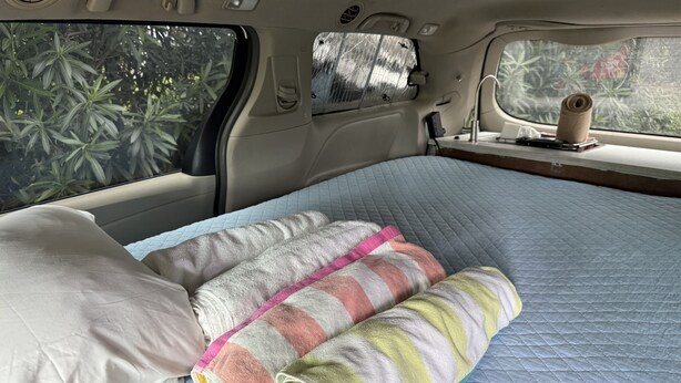 L’intérieur d’un véhicule aménagé avec un lit, des oreillers, un matelas matelassé, une fenêtre donnant sur la verdure et une petite table sur laquelle se trouvent un livre et un rouleau d’essuie-tout.