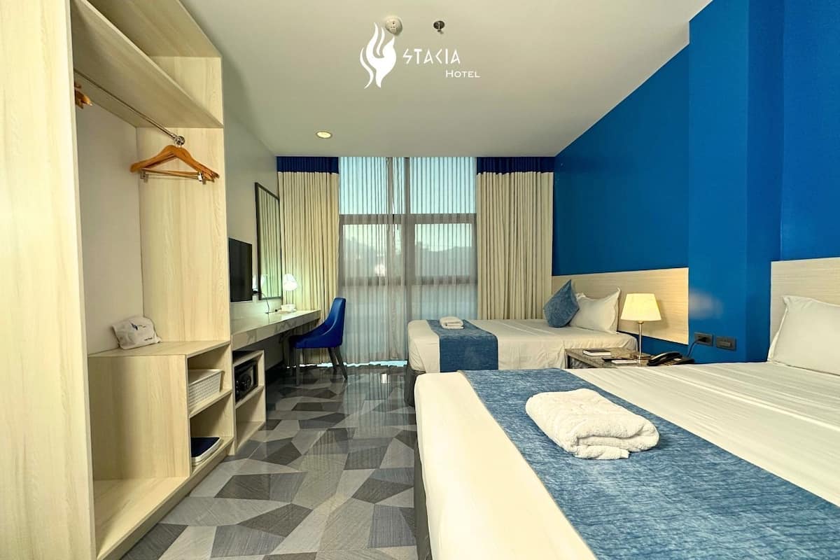 A hotel room with a desk, two beds, a wardrobe, and a view of the outdoors.