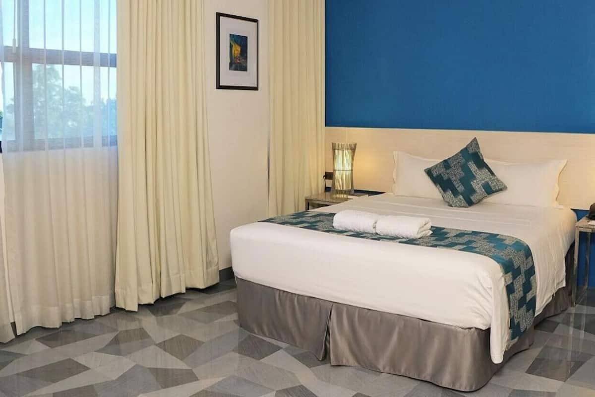A neatly made bed with a blue and white patterned bedspread, a white pillow, and a matching pillow on the headboard. There are two bedside tables with lamps, a framed picture on the wall, and a window with sheer curtains.