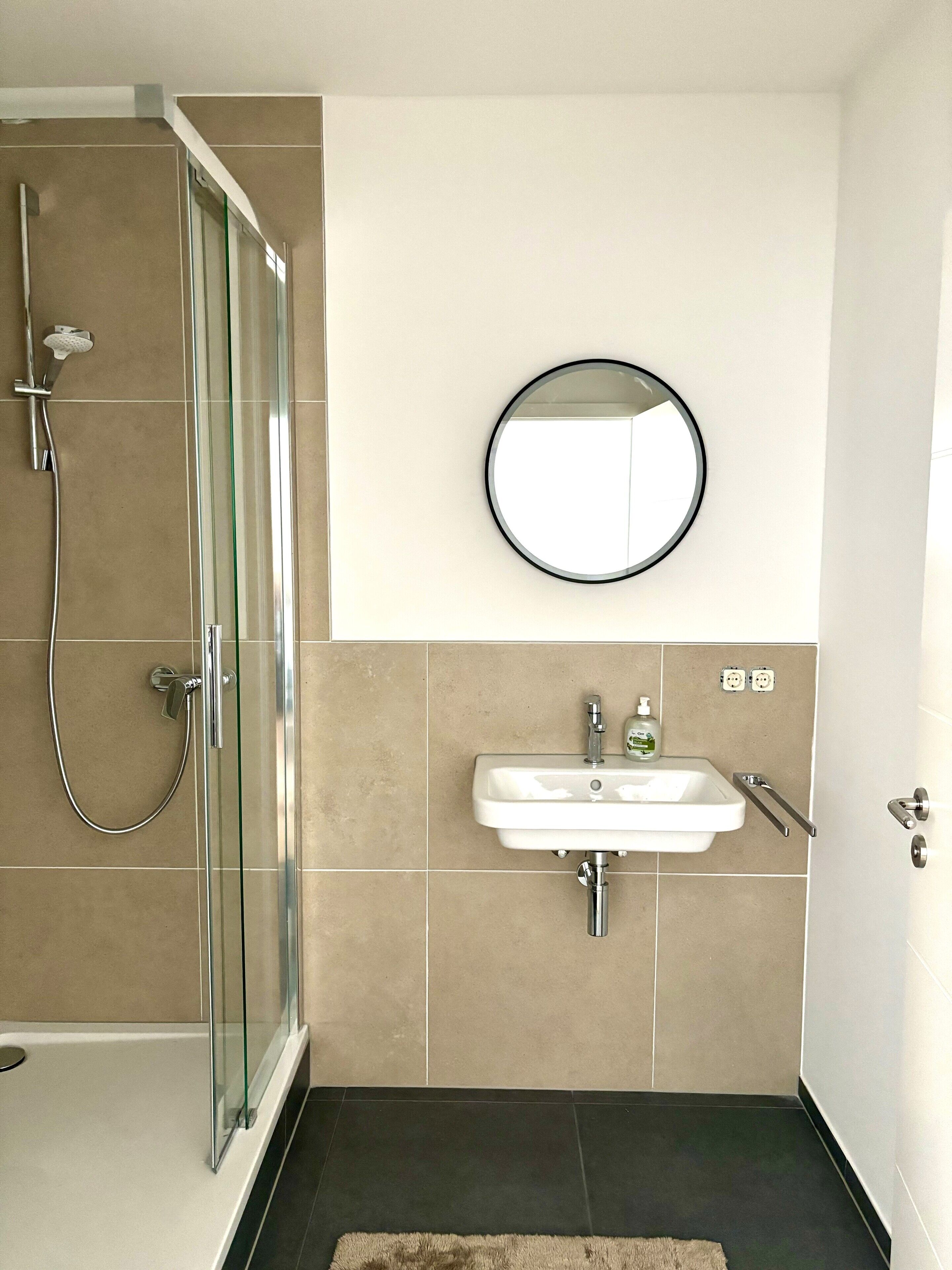 Ein modernes Badezimmer mit einer gläsernen Duschkabine, einem weißen Waschbecken mit Seifenspender und einem runden Spiegel an der Wand.