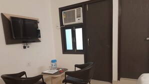A small room with a TV mounted on the wall, a window with an air conditioner, a desk with a chair, and a bed. - Hotel M A International (New Delhi)