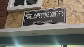 Exterior del Hotel White Stone Comforts, con un cartel con el nombre y la dirección del hotel, un gran ventanal con vista al interior y una puerta metálica en la entrada.