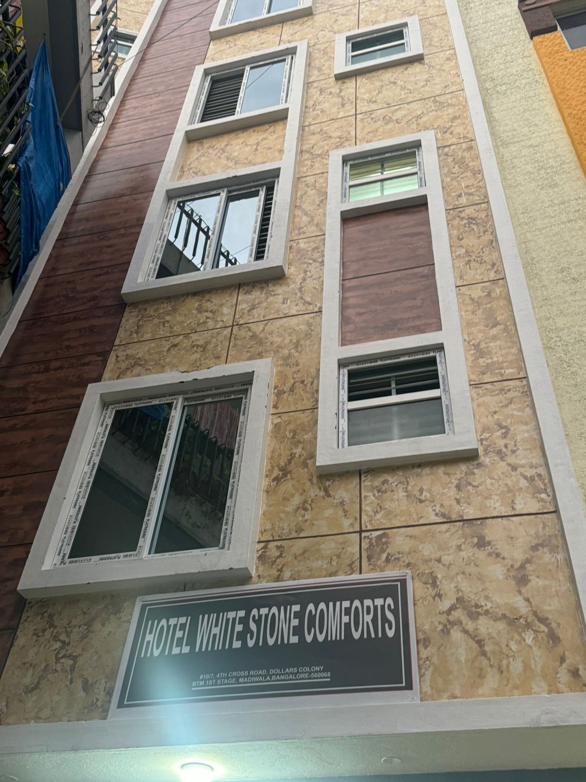 Fachada de hotel con múltiples ventanas, un letrero que dice "HOTEL WHITE STONE COMFORTS" y una vista parcial de un toldo azul a la izquierda.