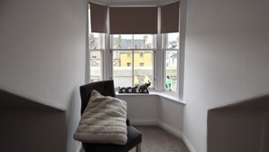 Une pièce d’angle avec une chaise noire et un plaid en fausse fourrure blanche, une grande fenêtre offrant une vue sur les immeubles et un store vénitien.