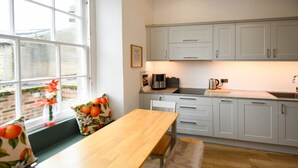 Une cuisine moderne avec des meubles de couleur gris clair, une table en bois et une fenêtre donnant sur un mur de briques et la verdure à l’extérieur.