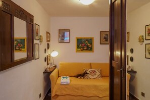 A cozy room with a yellow sofa, a small table with a lamp, and several framed artworks on the walls.