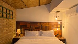 Un dormitorio con pared de ladrillo, cabecera de madera, cama con ropa blanca y mesita de noche con lámpara.