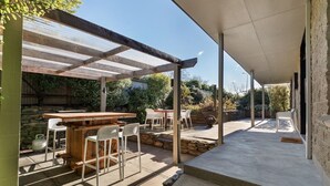 An outdoor patio area with a wooden bar and white chairs, surrounded by stone walls and greenery. - Large, spacious home great for large numbers (Cromwell)