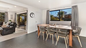 A modern living room with a dining area, featuring a sofa, a dining table with chairs, and a large window with curtains. - Large, spacious home great for large numbers (Cromwell)