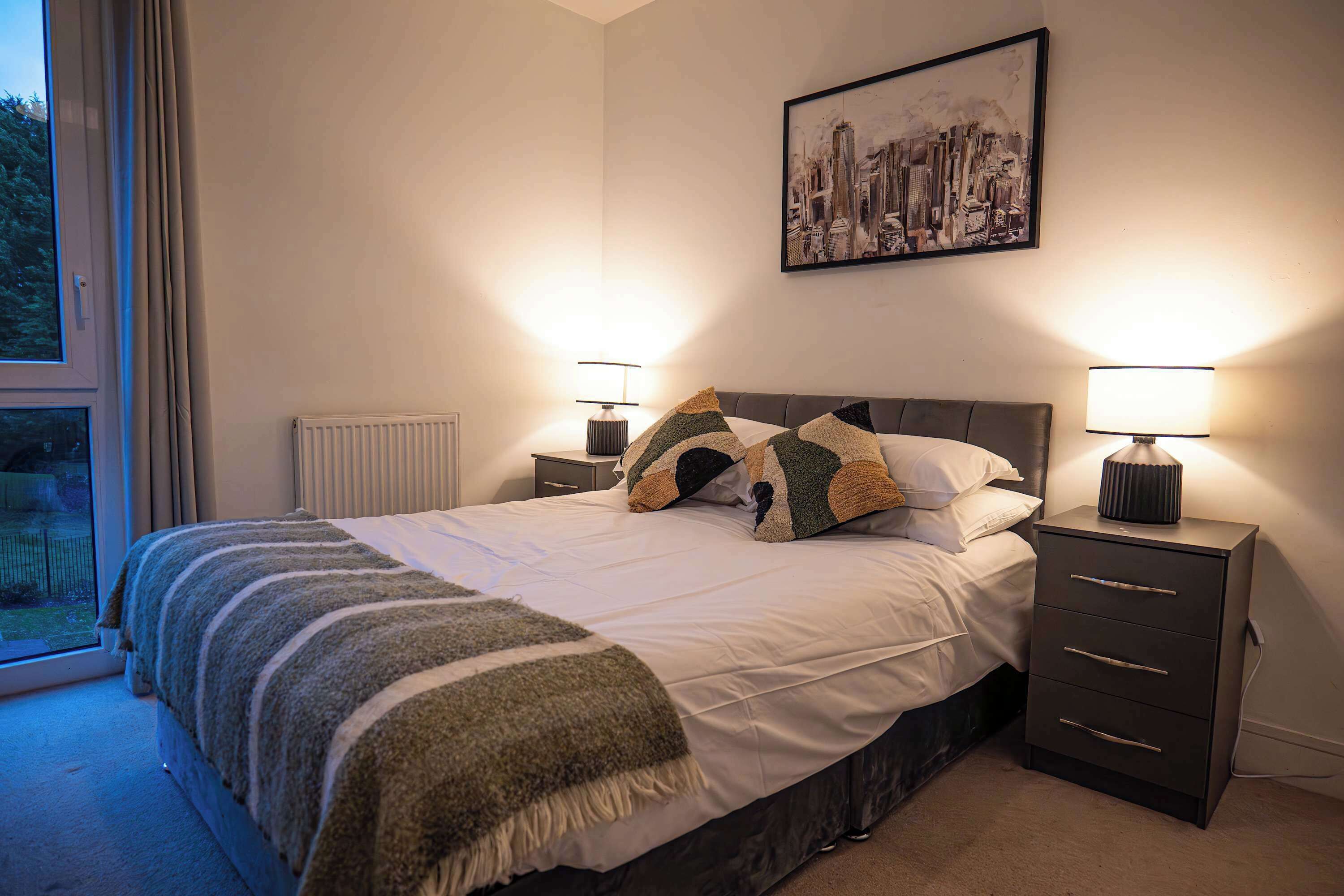 A bedroom with a bed, two bedside tables with lamps, a radiator, and a window with curtains.