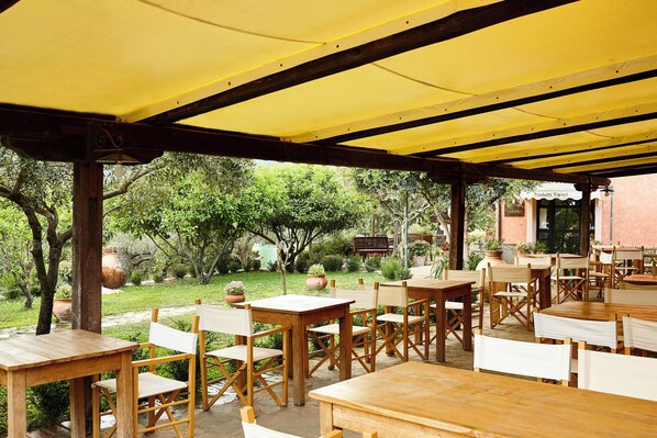 Un espace extérieur aménagé en terrasse, avec des tables et des chaises en bois, entouré de verdure et surmonté d’un auvent jaune.