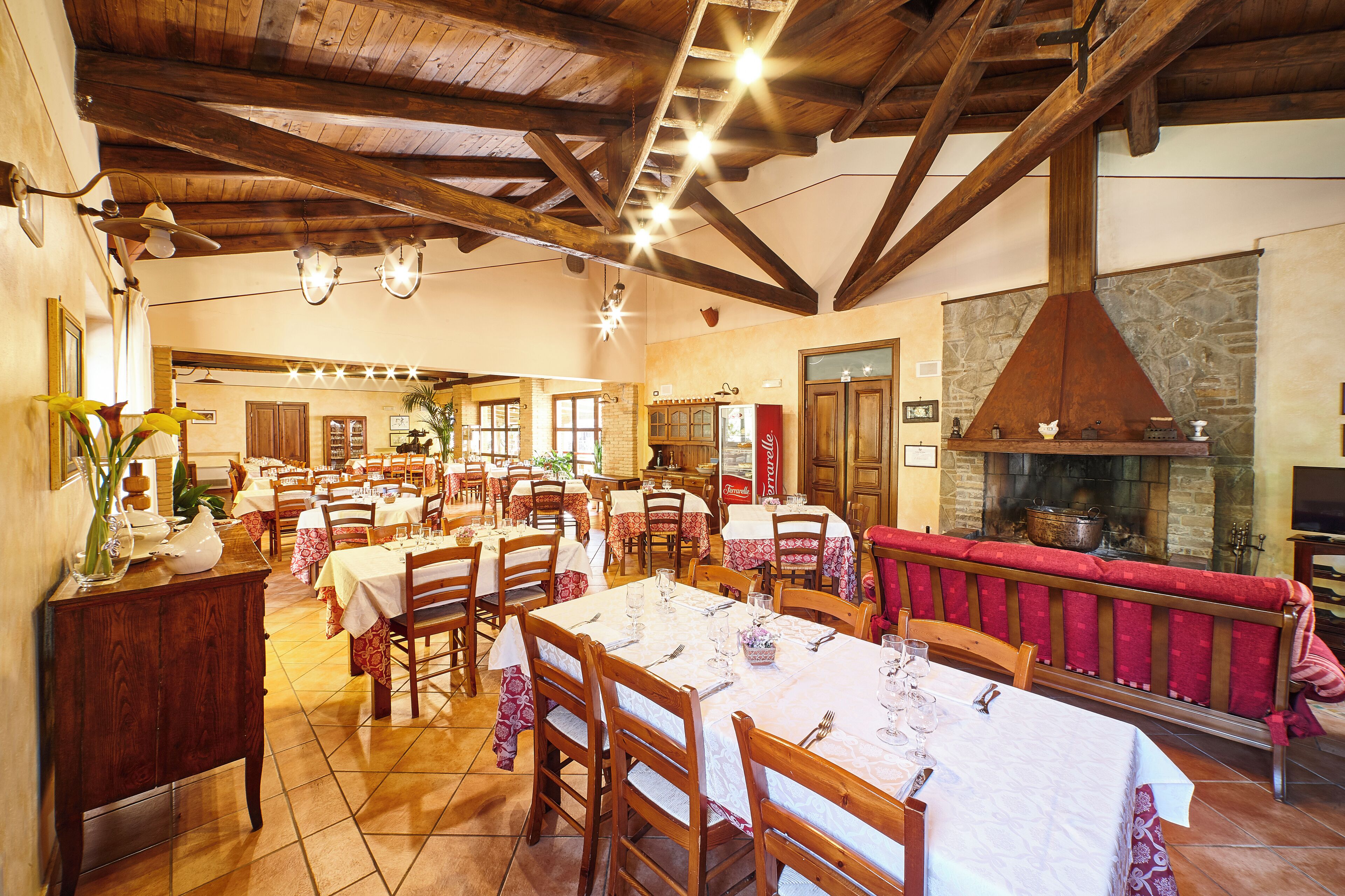 Un restaurante con vigas de madera, paredes de piedra y chimenea. Las mesas están dispuestas con manteles blancos y servilletas rojas.