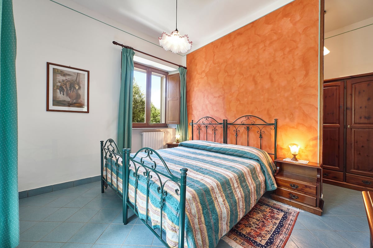 Een slaapkamer met een bed, nachtkastje, raam met gordijnen, radiator en een houten kledingkast.