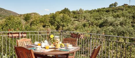 Una mesa redonda de madera con sillas en una terraza, rodeada por una barandilla de metal, con vistas a un paisaje montañoso con árboles y un edificio a lo lejos.