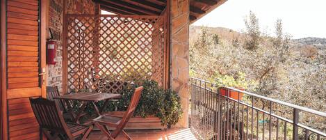 A wooden balcony with a table and chairs, a stone wall, and a view of a hilly landscape.