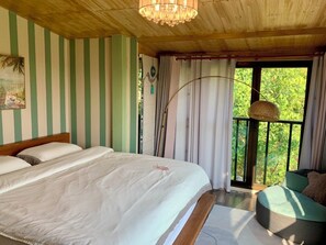 Une chambre avec un grand lit, un balcon agrémenté d’un fauteuil en osier et une vue sur la verdure.