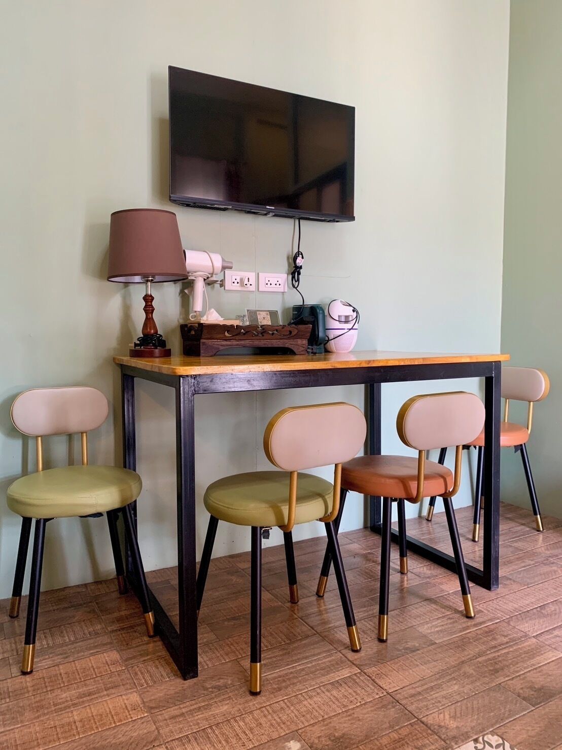 A small room with a wooden table, four chairs, a television mounted on the wall, and a lamp on the table.