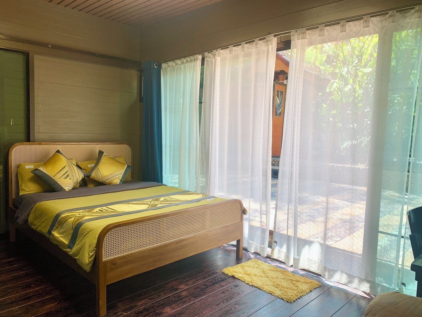 Une chambre avec un lit en bois, une literie jaune et une vue sur l’extérieur à travers des rideaux transparents.