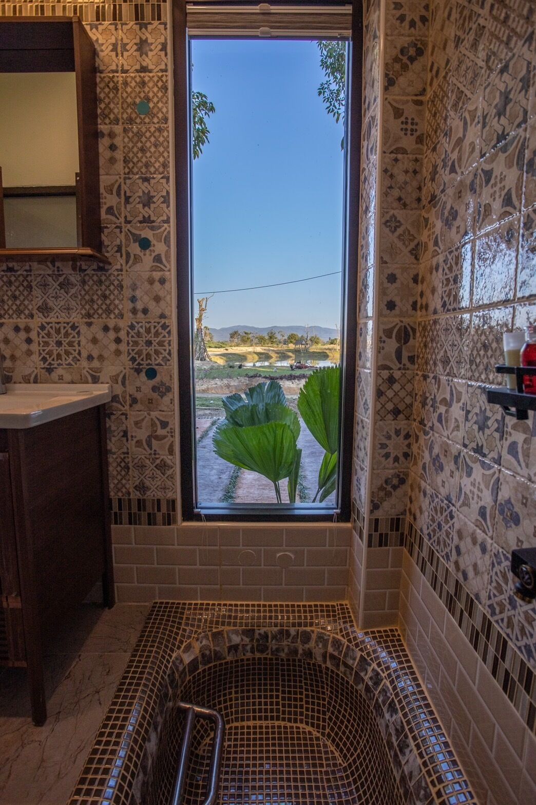 A bathroom with a large window, patterned tiles, a bathtub, and a wooden cabinet.