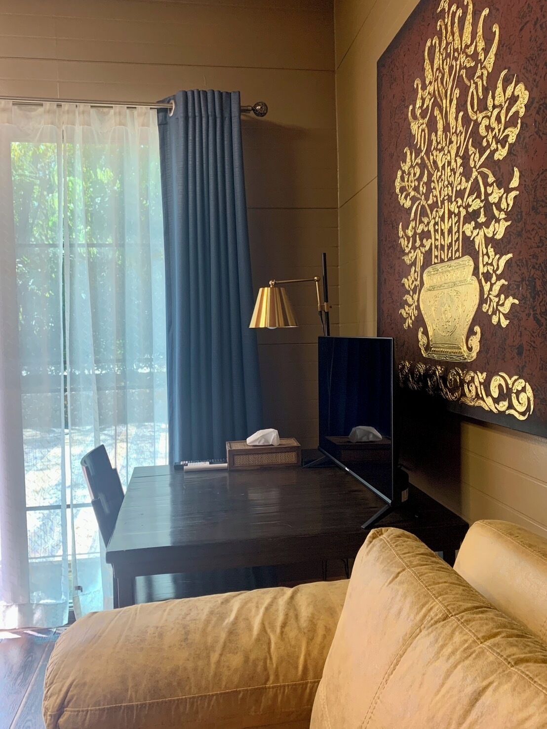 Una habitación con un escritorio de madera oscura, una cortina azul, una lámpara dorada y un sofá color beige.