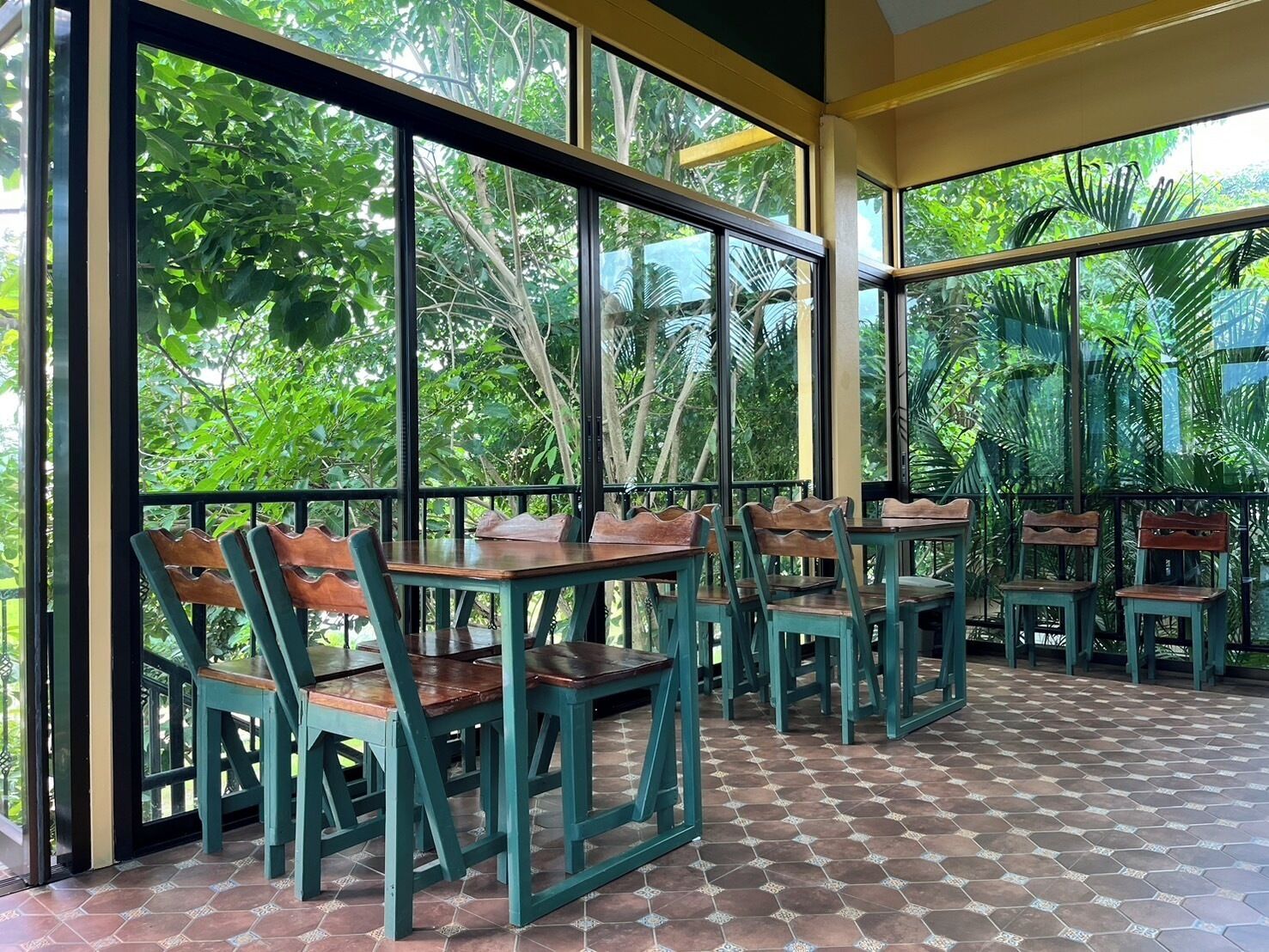 Un restaurant doté de grandes fenêtres offrant une vue sur une végétation luxuriante. Des tables et des chaises sont disposées pour accueillir les clients.