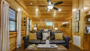 A cozy log cabin interior with a leather sofa, a ceiling fan, and a dining table with a tray. - Cozy cabin-Great Location (Hochatown)