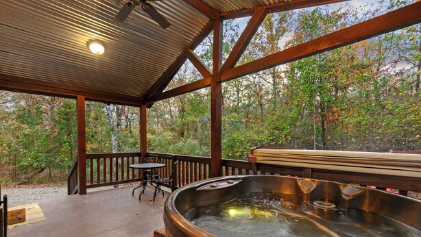 A wooden deck with a hot tub, a table, and chairs, overlooking a forested area. - Cozy cabin-Great Location (Hochatown)