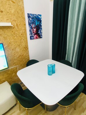 A room with a white table, green chairs, a painting on the wall, and a window with curtains.
