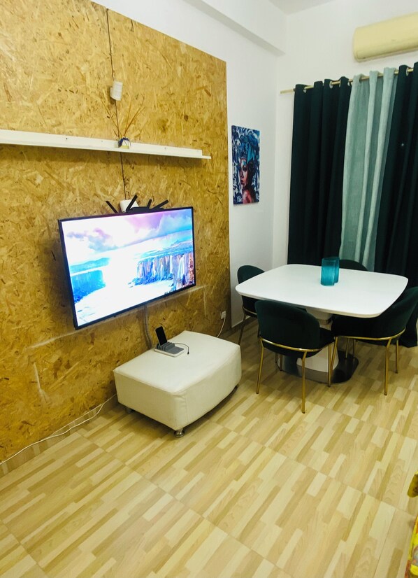 A modern living room with a flat-screen TV mounted on a wooden wall, a white ottoman with a charging device, a dining table with chairs, and a window with curtains.