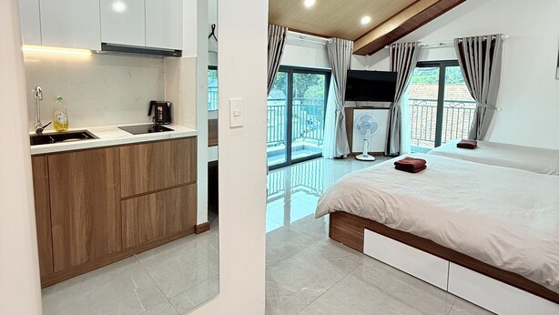 A modern bedroom with a large bed, a built-in desk, and a kitchen area with white cabinets and a wooden countertop. - Hasa Da Lat Hotel and Apartment (Xuan Huong - Da Lat)