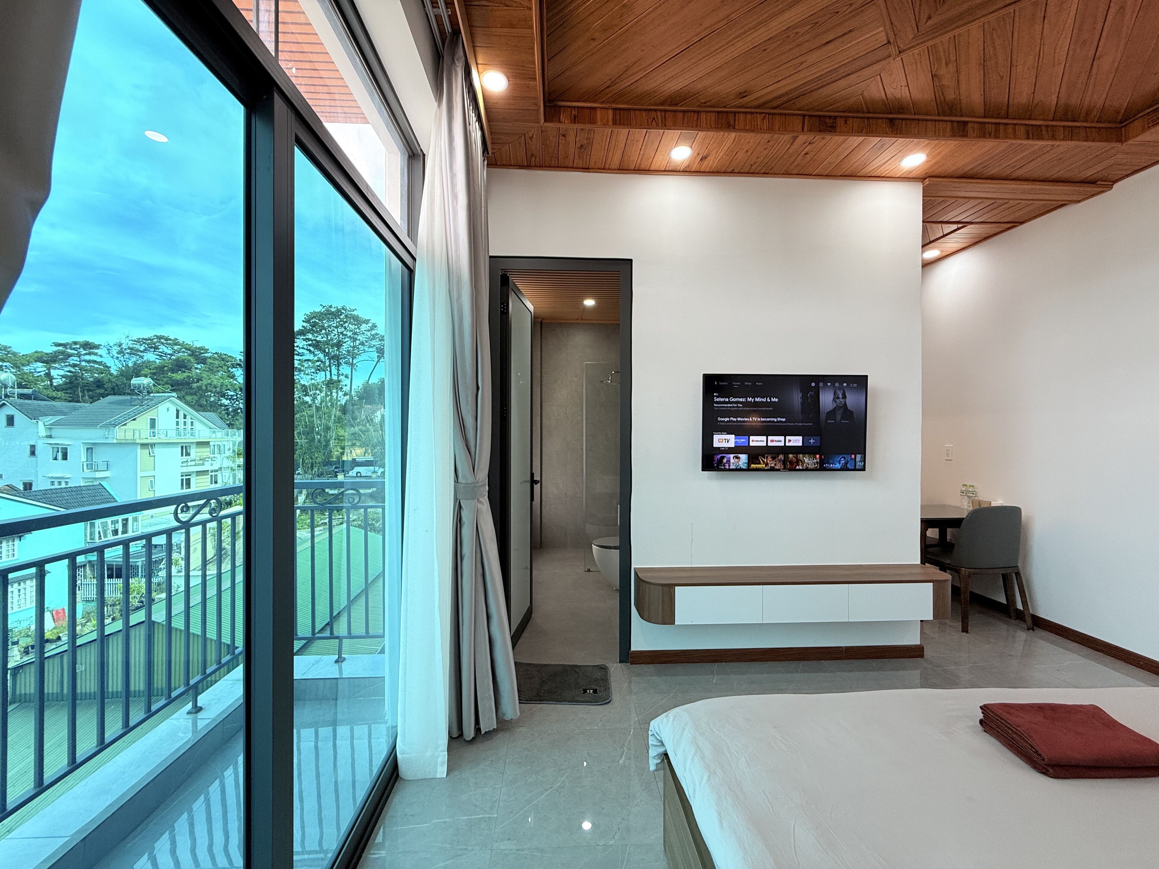 A modern hotel room with a large window, a flat-screen TV mounted on the wall, a wooden ceiling, and a minimalist design.
