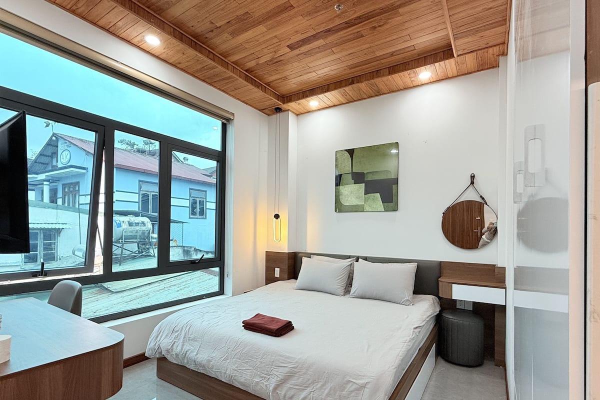 A modern bedroom with a large window, a bed, a bedside table, and a wooden ceiling.