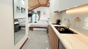 A compact hotel room with a kitchenette, a bed with a headboard, a small seating area, and a view through a window with curtains. - Hasa Da Lat Hotel and Apartment (Xuan Huong - Da Lat)