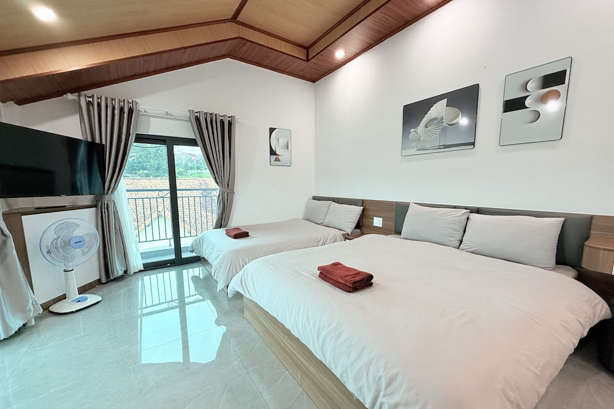 A hotel room with two beds, a large window with curtains, a ceiling fan, and artwork on the walls.