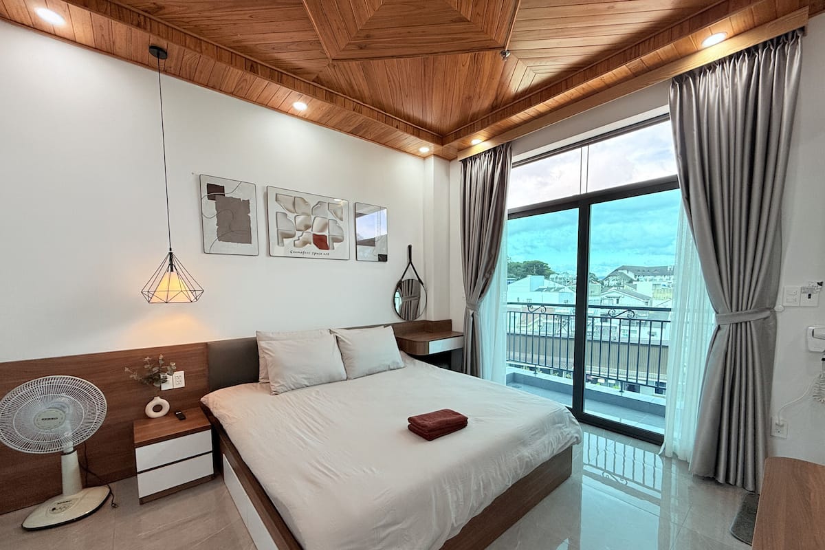 A modern bedroom with a wooden ceiling, a large bed with white bedding, a bedside table with a fan, and a view of the city through a large window.